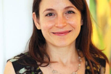 A portrait of a smiling woman with long brown hair, wearing a patterned dress and pearl necklace, featured in Oregon's 1859 Magazine, highlighting Oregon's culture, arts, and lifestyle.