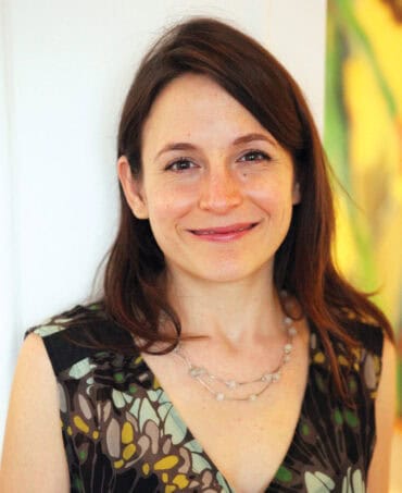 A portrait of a smiling woman with long brown hair, wearing a patterned dress and pearl necklace, featured in Oregon's 1859 Magazine, highlighting Oregon's culture, arts, and lifestyle.