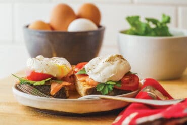 Close-up of poached eggs on toast with fresh vegetables, including tomatoes and greens, served on a rustic plate with a bowl of eggs and a salad in the background.