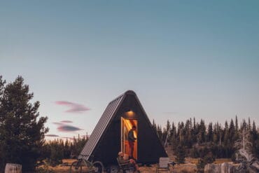 A cozy A-frame cabin in Oregon's wilderness during sunset, surrounded by trees and open grassy fields, with a person sitting on a bench outside, capturing the serene mountain landscape.