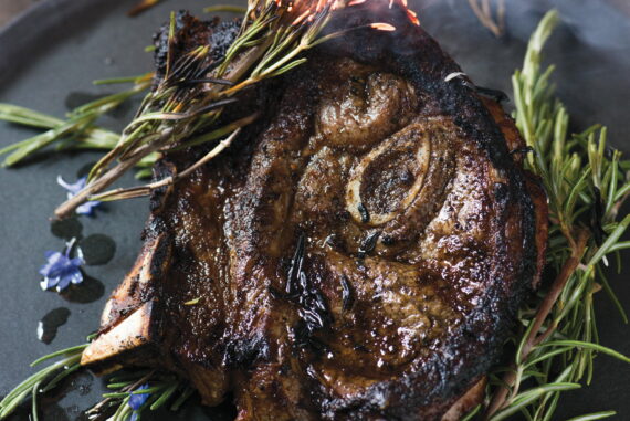 Juicy roasted lamb leg garnished with fresh rosemary and edible flowers, served on a black platter. Perfect for gourmet dining and special occasions.