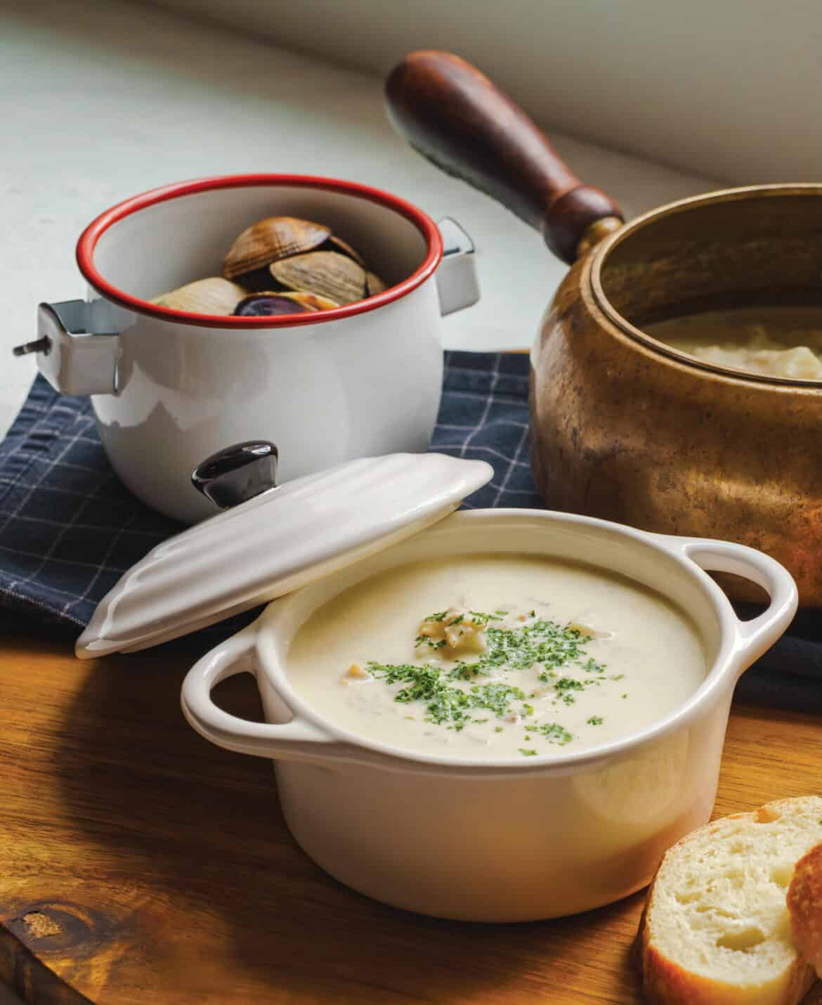 A bowl of creamy clam chowder garnished with herbs, accompanied by fresh bread and a pot of clams, showcasing a comforting seafood dish from Oregon.