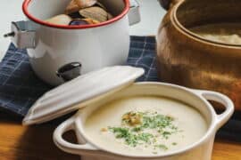 A bowl of creamy clam chowder garnished with herbs, accompanied by fresh bread and a pot of clams, showcasing a comforting seafood dish from Oregon.