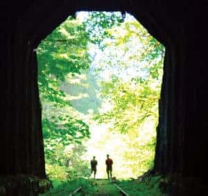 Scenic view of two hikers standing at the entrance of a tunnel surrounded by lush green trees, highlighting the Salmonberry Trail development in Oregon.